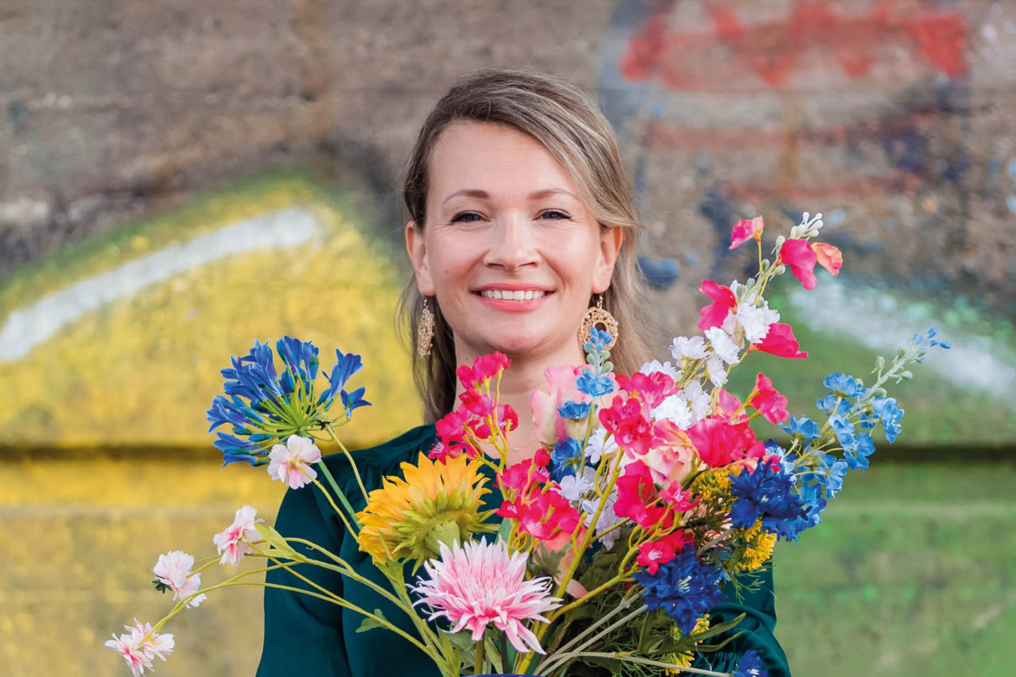 Cocky Drost is een vlotte vrouw en lacht in de camera. In haar handen heeft ze een grote bos bloemen met verschillende kleuren. Ze staat voor een muur met graffiti.
